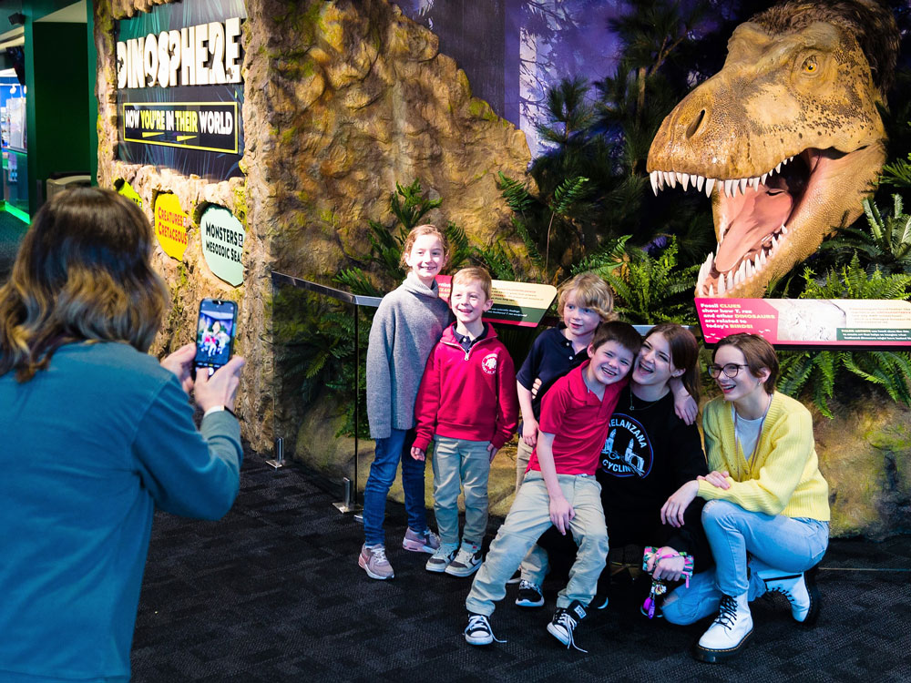 Family posting for a photo in front of a sculpture of a feathered Tyrannosaurus rex head at the entrance to the Dinosphere exhibit.